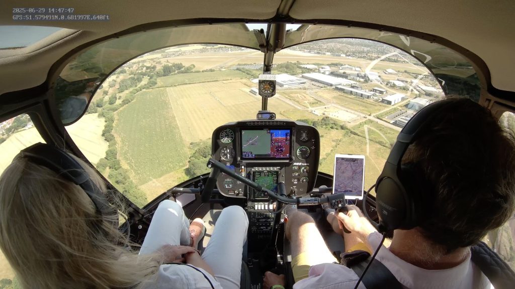 View from a helicopter cockpit flying over fields with Helipaddy GPS and map navigation tools in use.