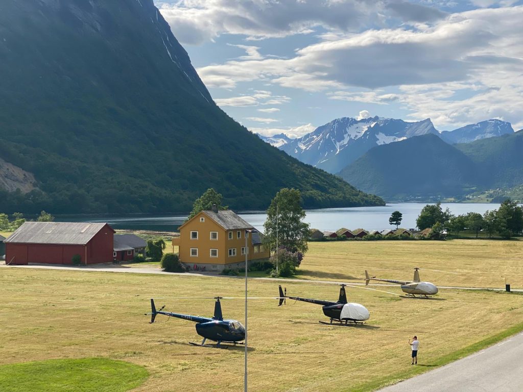 Helipaddy helicopters parked on scenic field with mountain and lake views, near a vibrant yellow house.