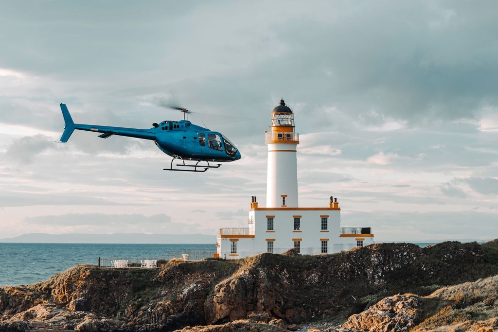 A blue helicopter flies near a lighthouse on a rocky coast, featured on Helipaddy.
