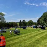 Helipaddy event with helicopters parked on a lush green field under a clear blue sky, attended by people in bright vests.