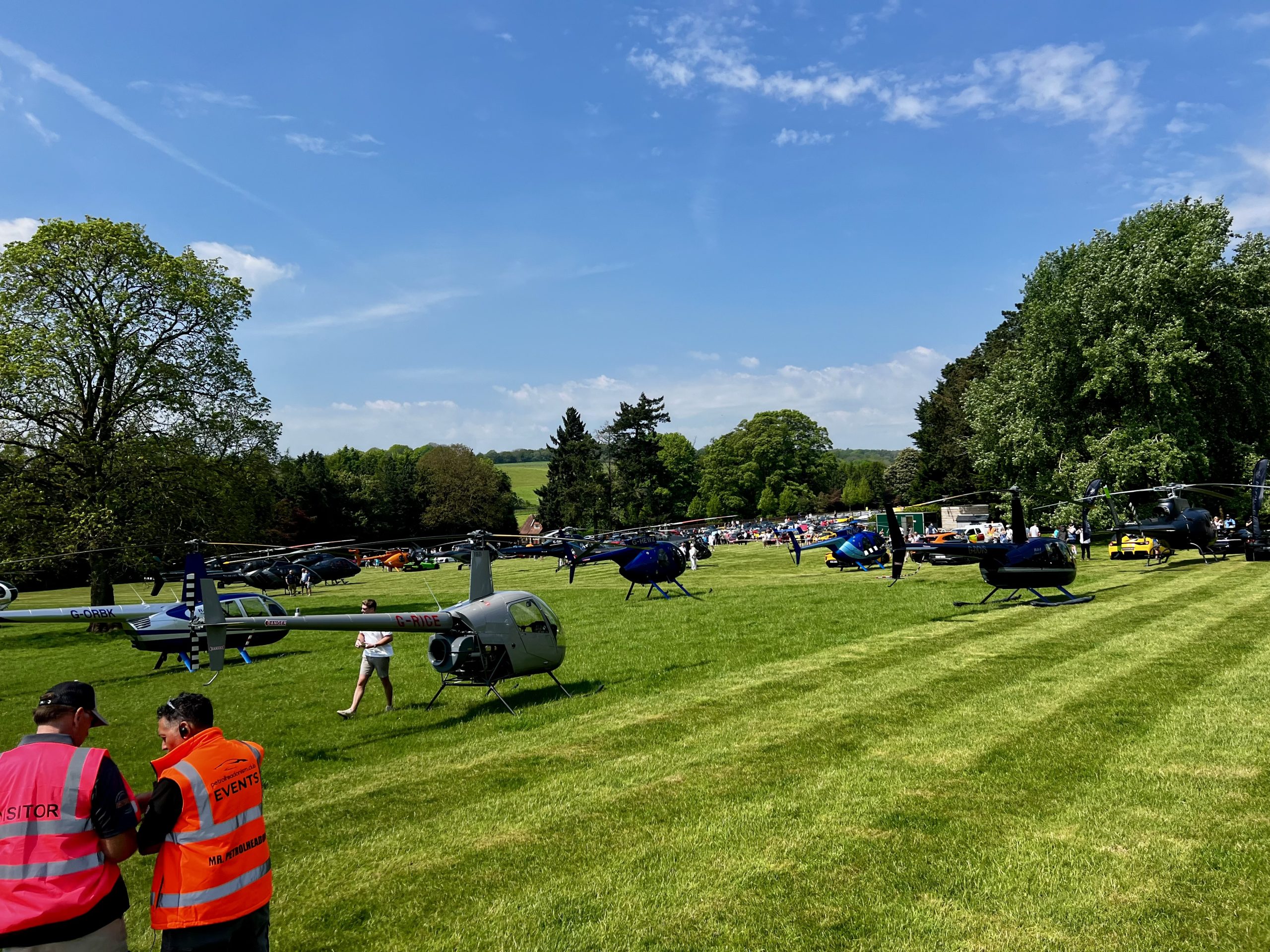 Helipaddy event with helicopters parked on a lush green field under a clear blue sky, attended by people in bright vests.